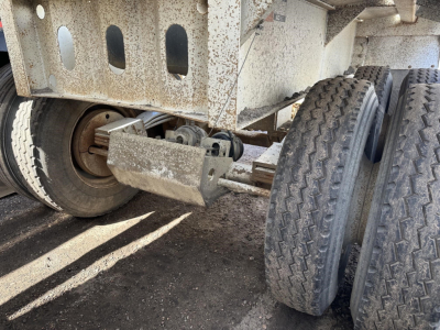 Trailer axle and wheel assembly of a dump trailer showing dual rear tires and rusty steel frame with hitch components.