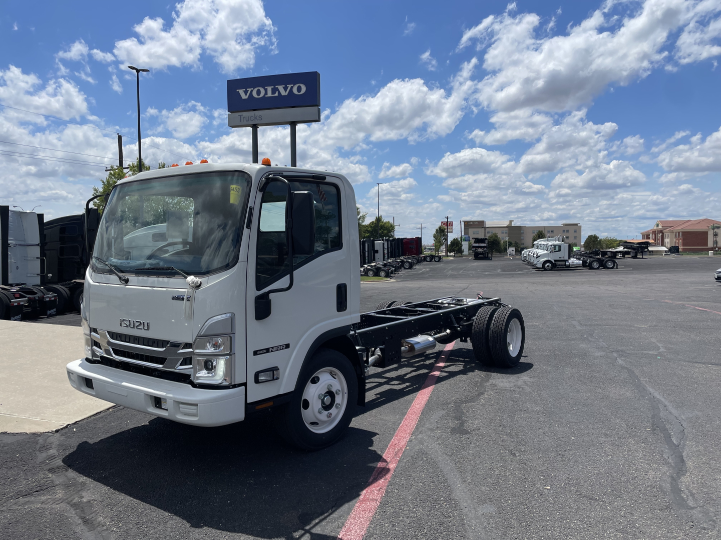 Isuzu NRR cab and chassis truck in a dealership lot showing front view with chassis back, Isuzu logo, and Volvo sign in background.