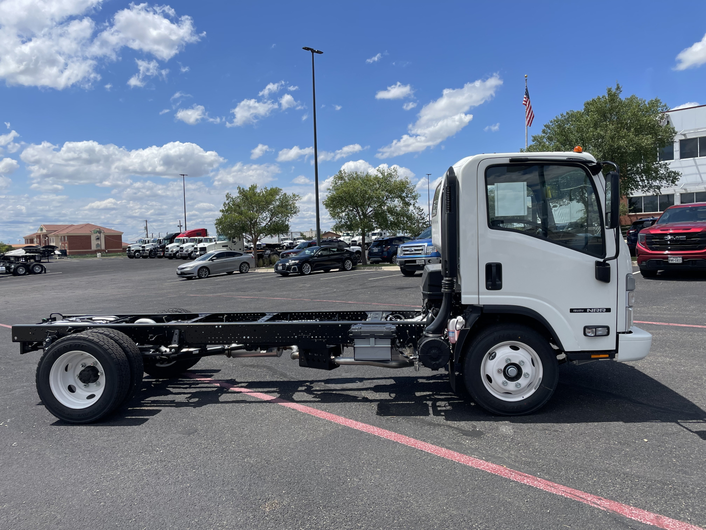Isuzu NRR cab and chassis truck in white, side view, showing chassis frame and front cab with Isuzu badge. Clear blue sky with scattered clouds.