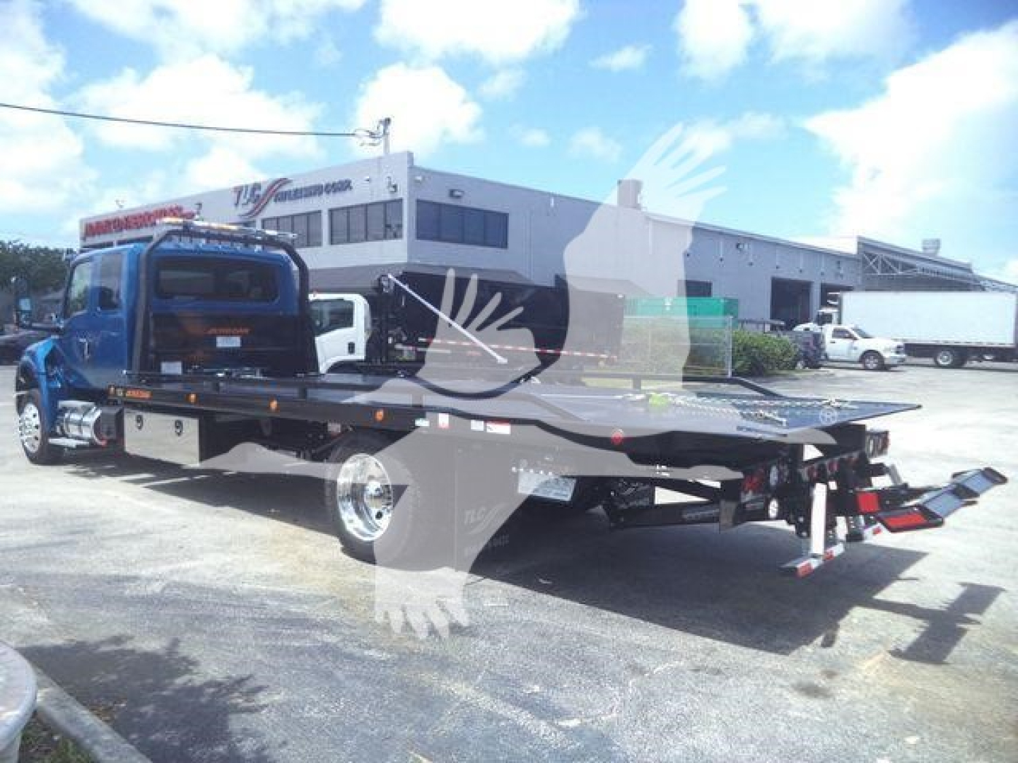 Flatbed tow truck with blue cab and long black flatbed, rear view showing hydraulic wheel-lift, on lot with dealership building in background.
