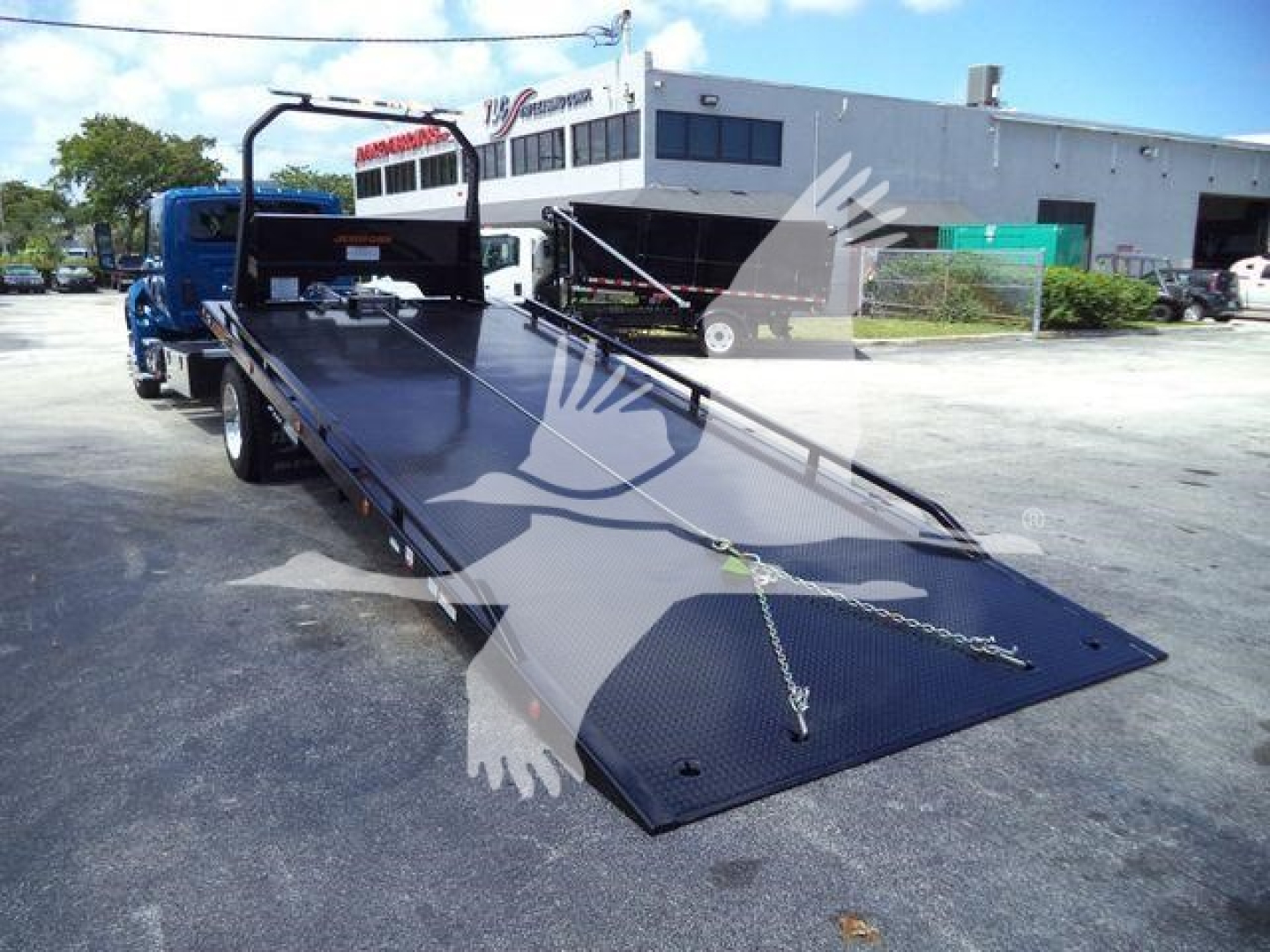 Flatbed tow truck with long black metal platform in a parking lot, chain tie-downs at the front, ramp extended downward, rear wheels visible, white building in background.