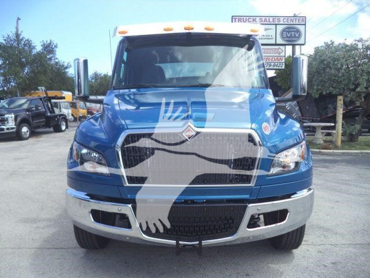 Front view of a blue 2027 International MV truck showing large grille, chrome bumper, and side mirrors. Visible text includes stock sign and dealership background.