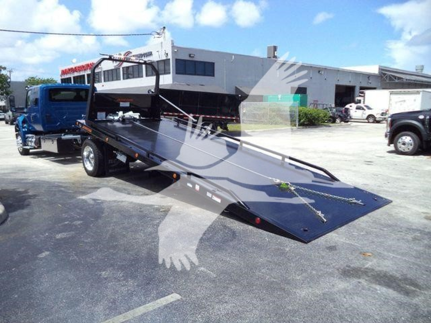 Flatbed tow truck loading ramp on a blue truck with a white International MV roll-back unit in an industrial lot.