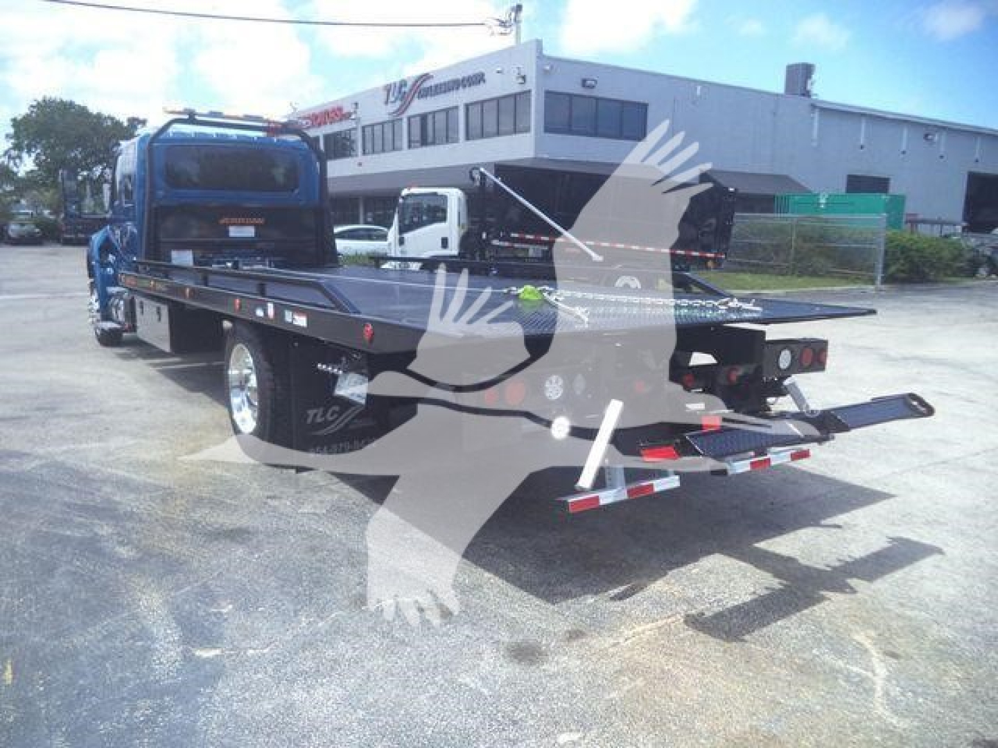 Flatbed tow truck trailer with blue cab, rear view showing extended steel bed and ramps, International MV stock image front lot