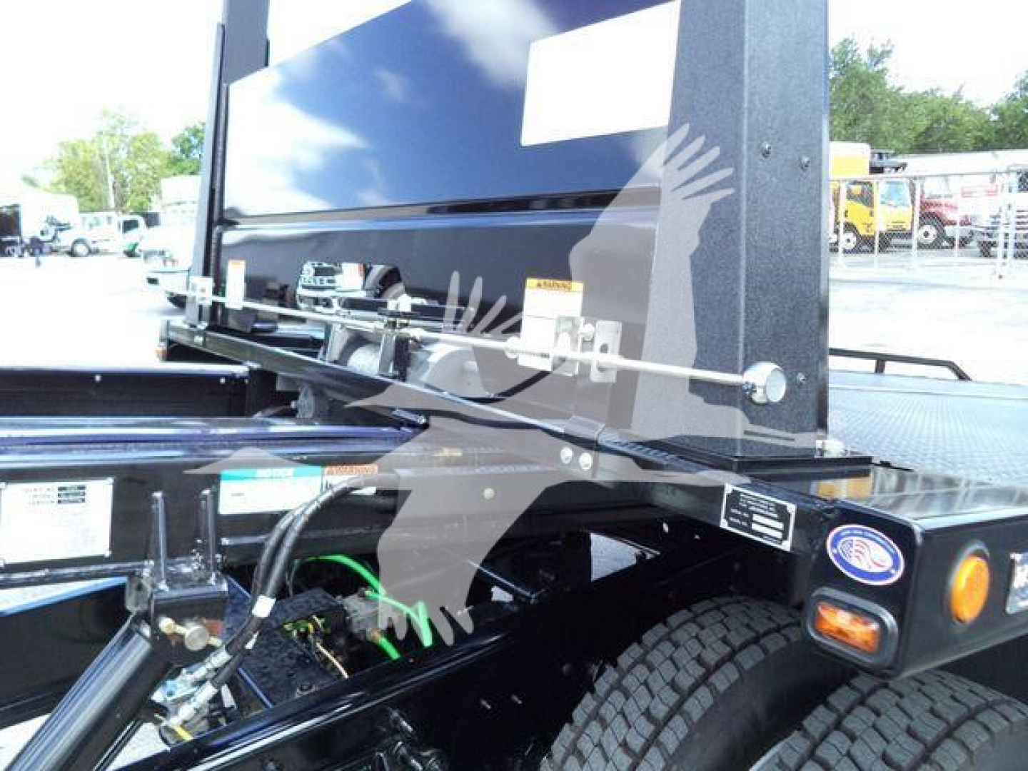Truck bed area showing a hydraulic rollback system with exposed hoses and a green hydraulic line, chassis components, and a black metal framework near the rear wheel.