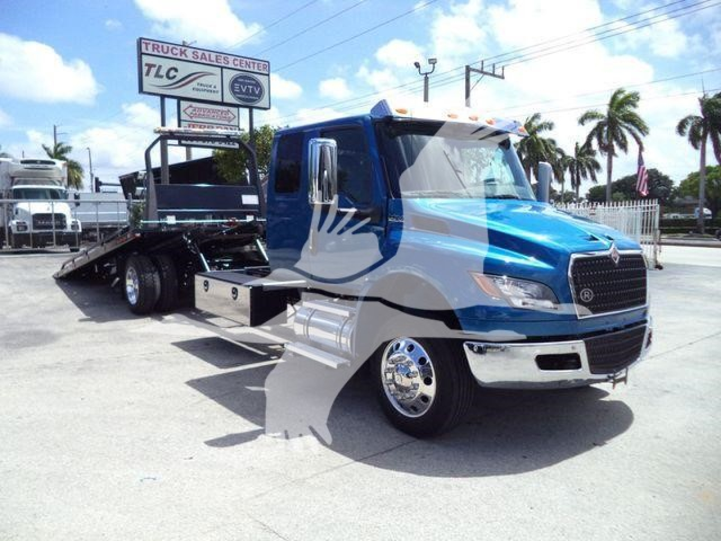 Blue International MV wrecker truck in a dealership lot, front chrome bumper, side chrome fuel tanks, rollback bed visible in background, palm trees and signboard in distance.