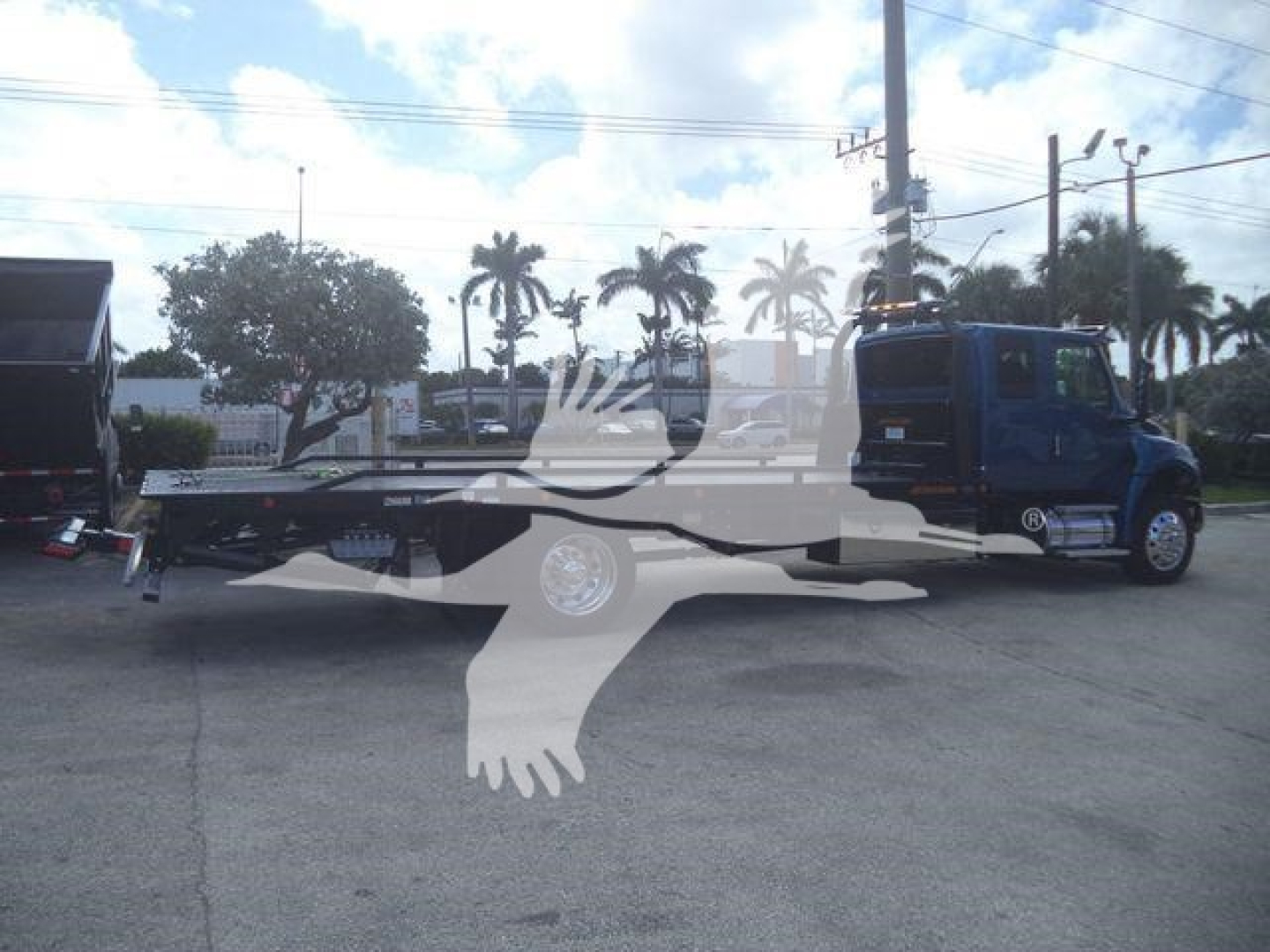 Truck body showing a blue International MV rollback configuration with long flatbed and wheel, side view, palm trees in background.