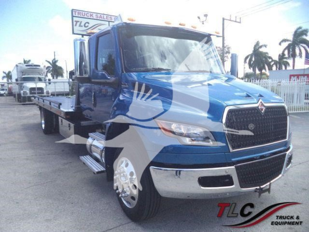 Truck with blue cab and flatbed, side view showing chrome bumper and large grille. Visible text on license area and dealership sign in background; no legible model numbers.