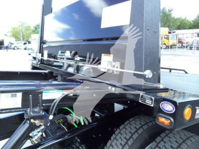 Truck bed area showing a hydraulic rollback system with exposed hoses and a green hydraulic line, chassis components, and a black metal framework near the rear wheel.
