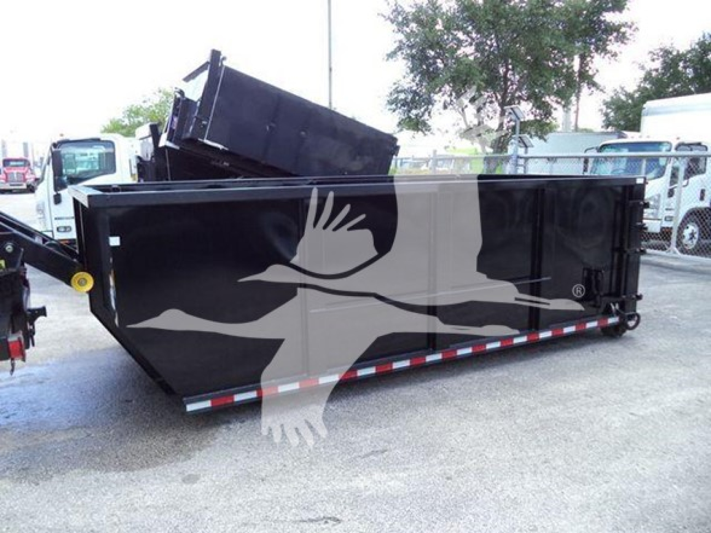 Flatbed truck dump body in a lot with a black roll-off dumpster in front; no legible brand text or serial numbers visible on the dumpster.