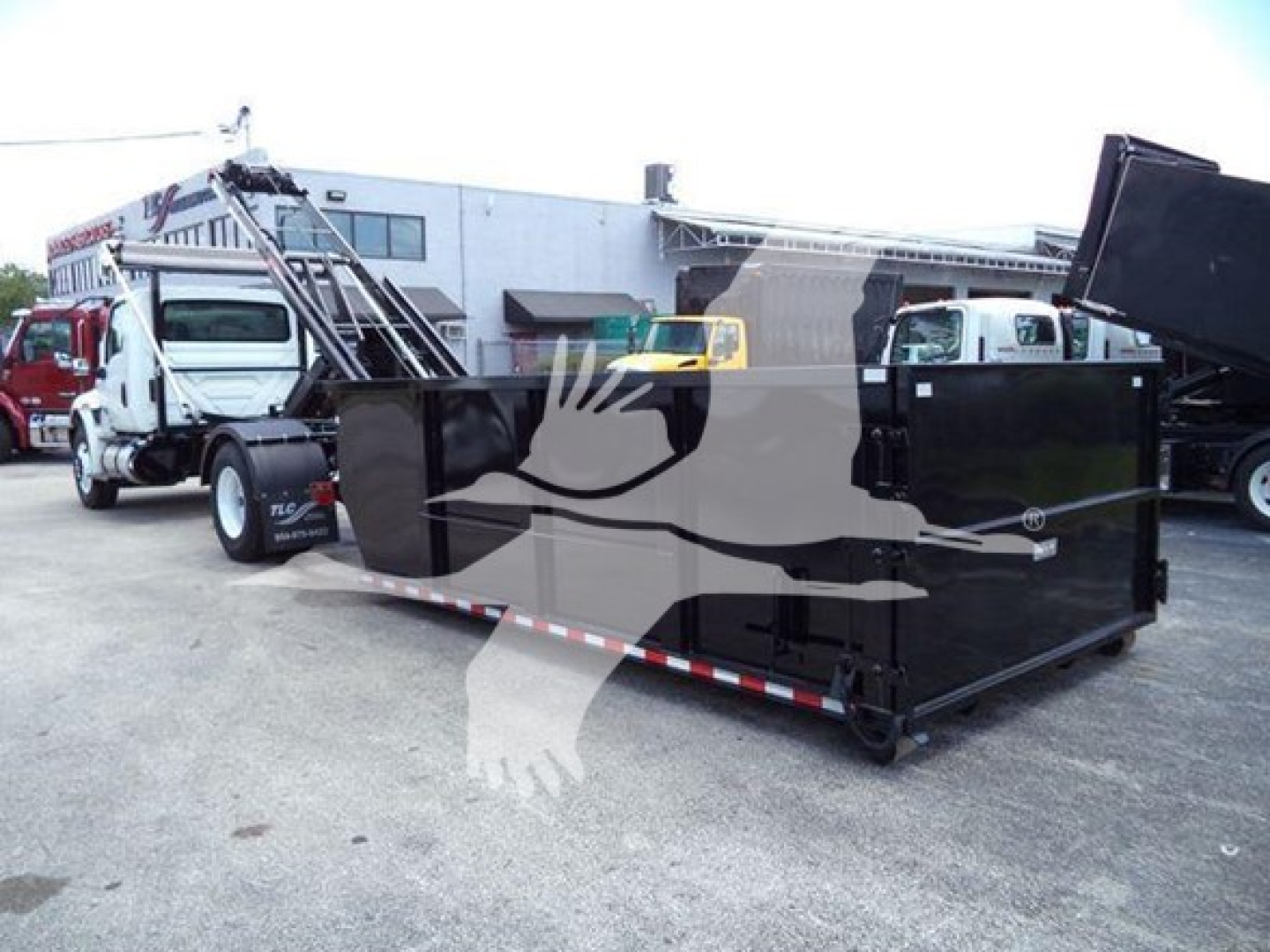 Dump truck body and trailer in a lot with a black roll-off dumpster container in foreground; faint text on building signs and trucks in background.
