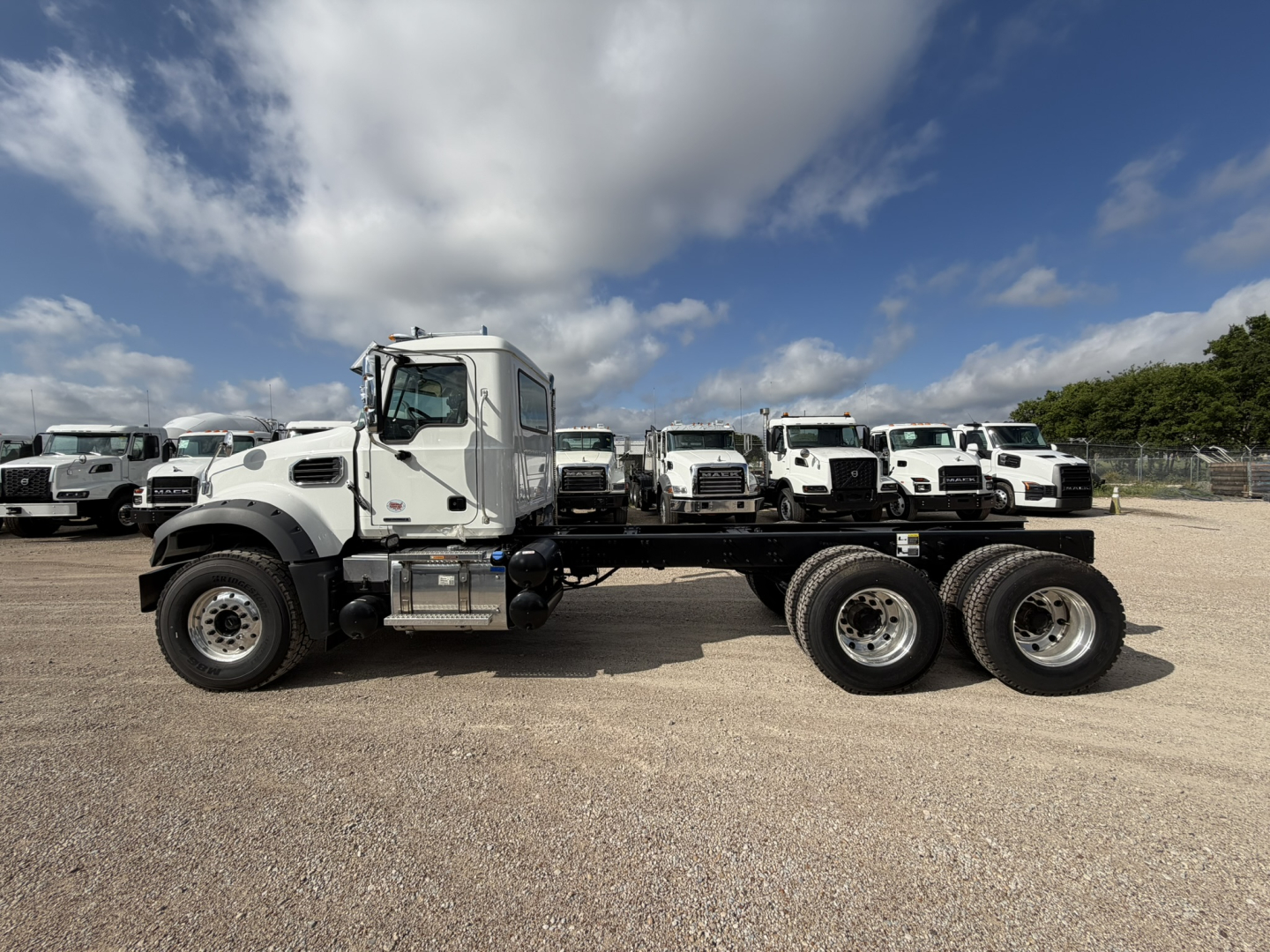 Truck chassis showing front cab and triple-axle rear with white Mack GR64F cab and chassis, parked in a lot with other trucks.