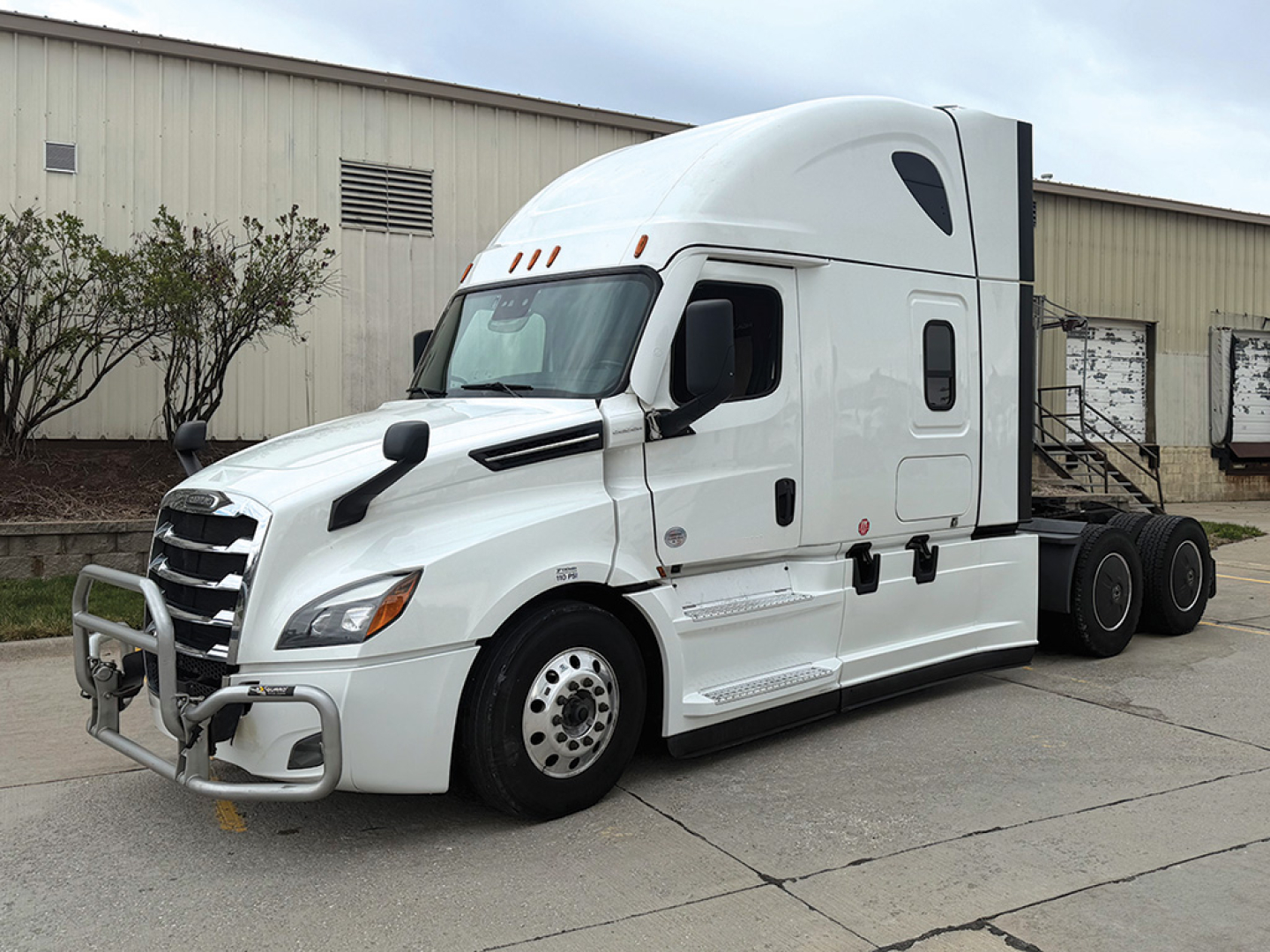 2022 Freightliner Cascadia 126 conventional sleeper tractor in white, side view with front bull bar and aerodynamic mirrors, parked on pavement beside a beige industrial building.