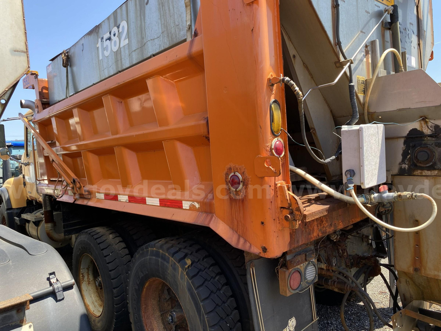 Dump truck bed on a 2005 Mack CV713 with orange cargo box and dual rear tires, side view showing tail lights and hydraulic lines, stock number and stickered identifiers.
