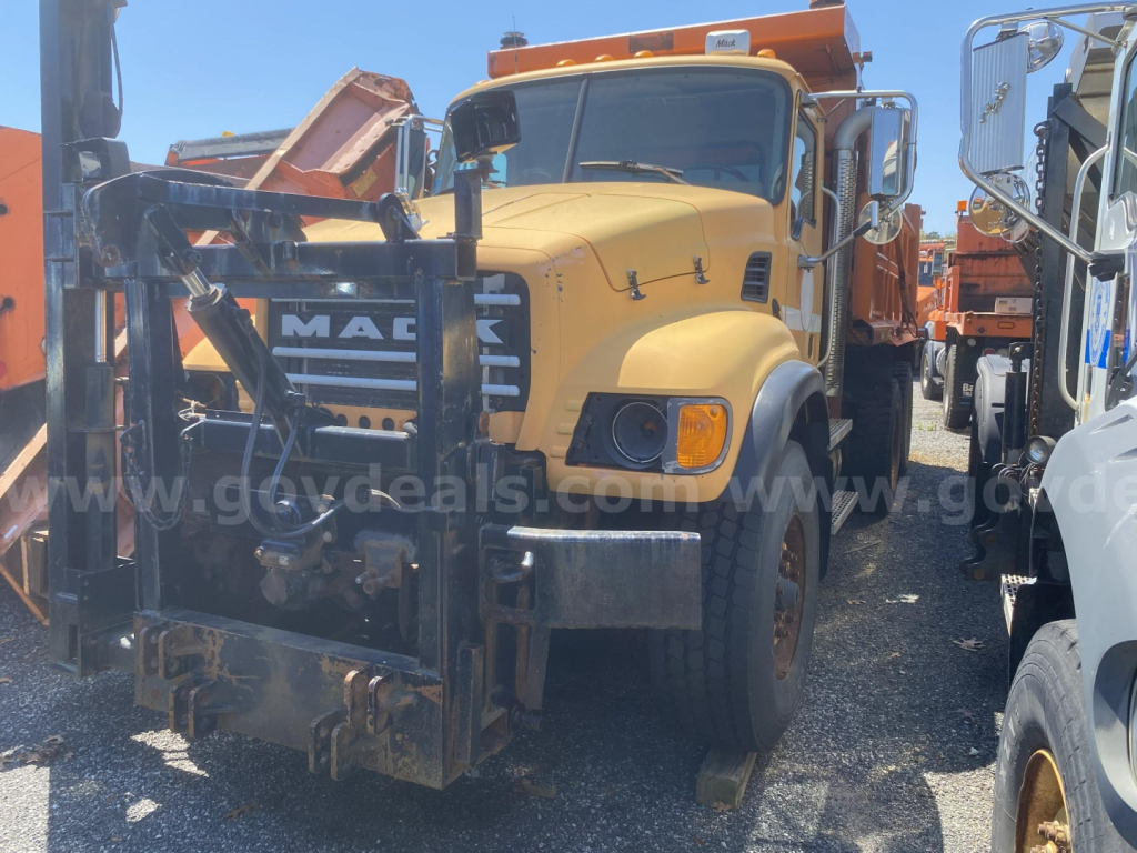 Mack CV713 dump truck front view showing front mount forklift/bale attachment and yellow cab with black grille and headlights.