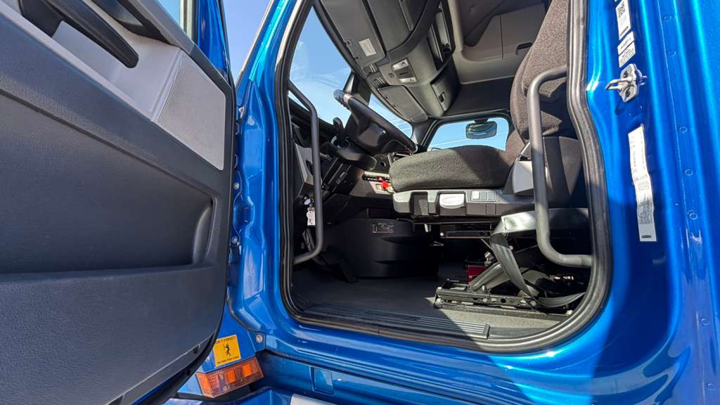 Cabin interior of a blue Freightliner truck with open driver door, view of seat and steering wheel from the doorway.