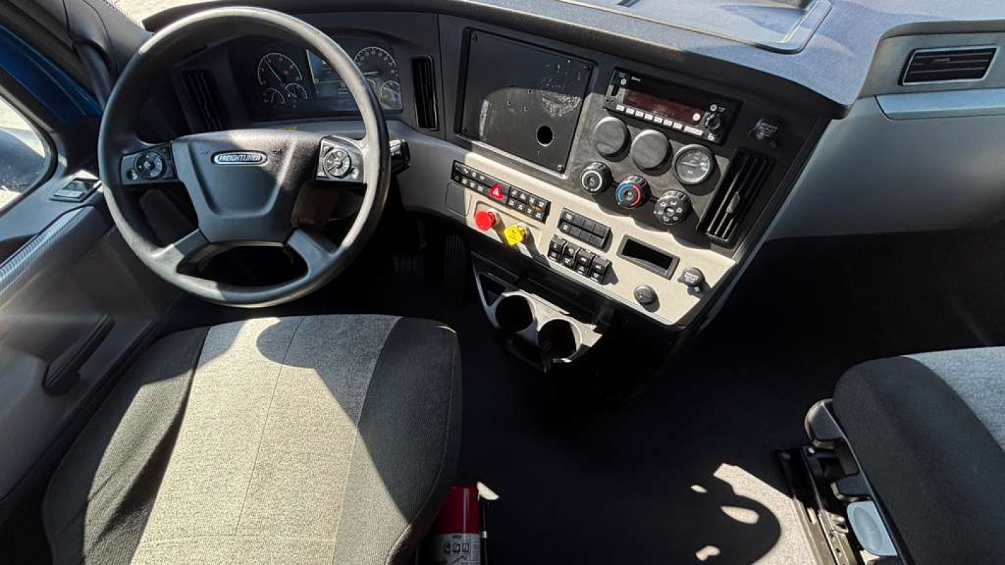 Cab interior of Freightliner truck showing steering wheel dashboard controls and center console with gauges and knobs.
