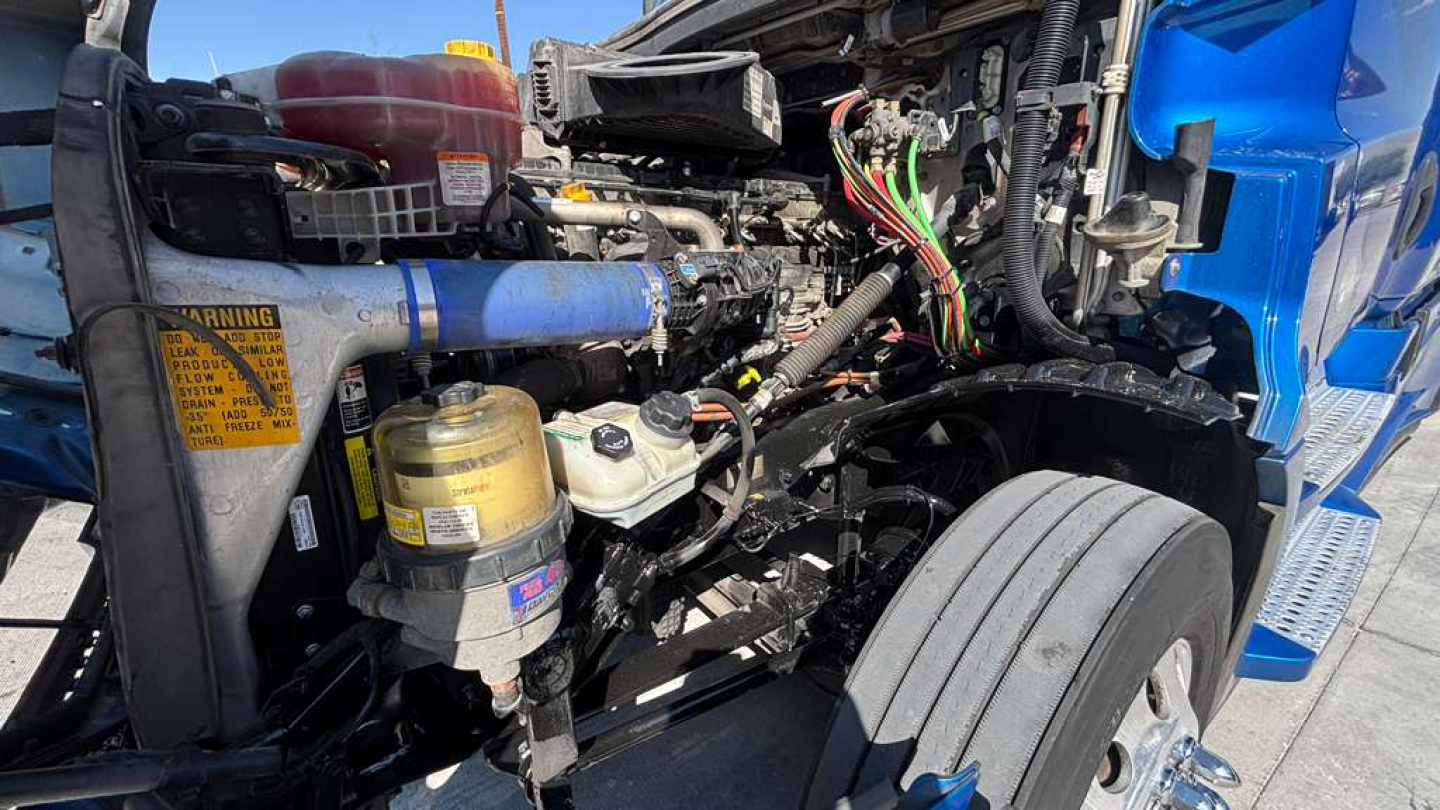 Truck engine bay of a Freightliner with exposed hoses, reservoir caps, and wiring harnesses visible.