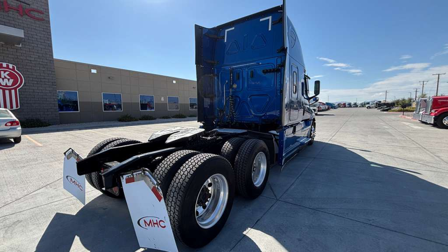 Truck in a dealership lot showing rear and side of a blue Freightliner cab with fifth wheel and dual rear axles, MHC logo on mud flaps, and dealership building in background.