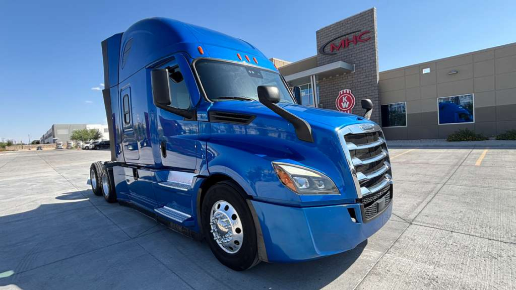 Blue Freightliner semi-truck tractor in a dealership lot, front-left angle, with MHC building in background.
