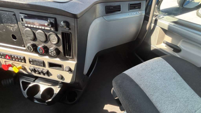 Dashboard and center console of a Freightliner truck showing gauges, knobs, vents, cup holders, and passenger seat. Visible labels include an emblem reading Freightliner and various control markings.
