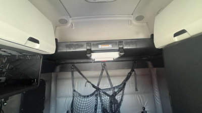 Truck cab interior showing sleeper area with overhead storage cabinets and window shade; a hanging net/strap on the back wall with metal bracket and warning label.