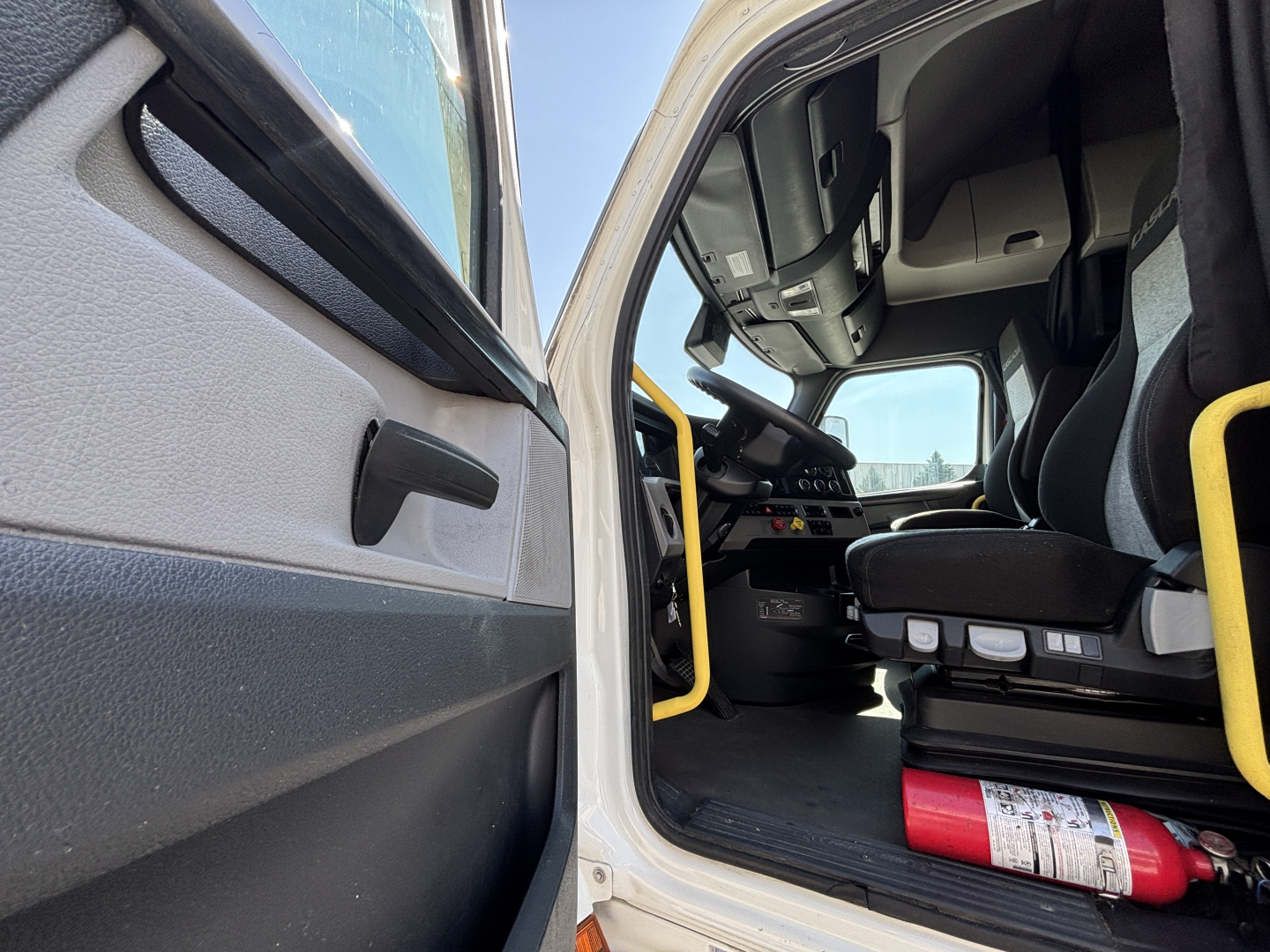 Cab interior of a Freightliner truck with open door, driver's seat visible, steering wheel, dashboard, and a red fire extinguisher on the floor.