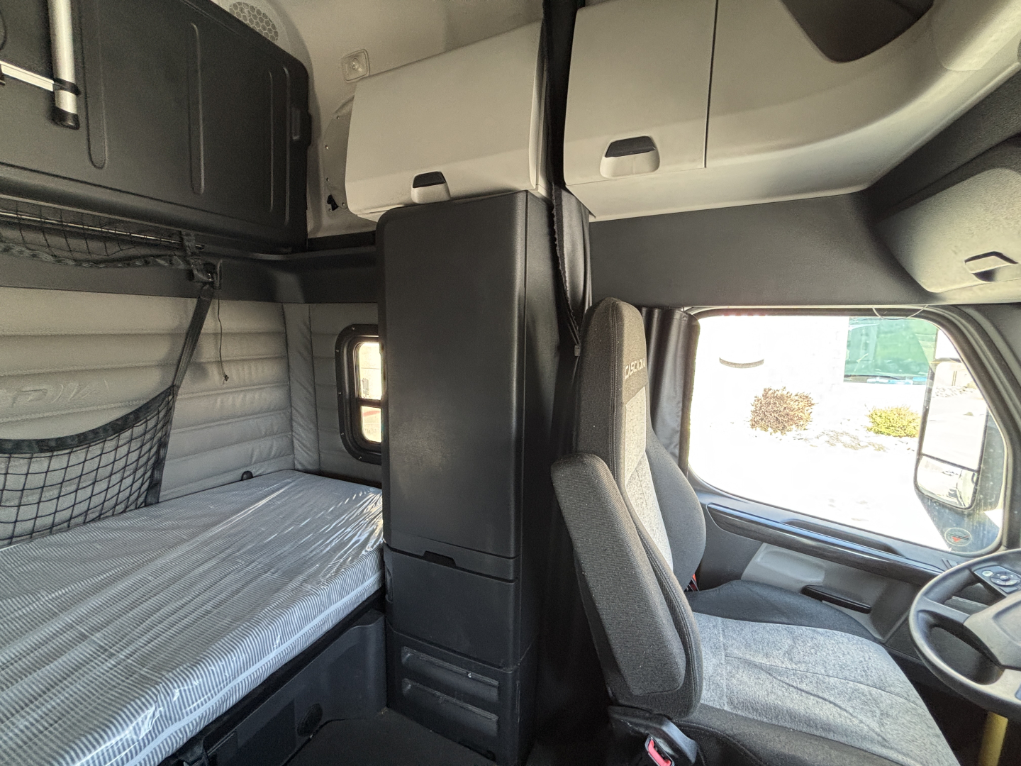 Cab interior of a Freightliner sleeper cab showing driver seat, overhead sleeper bunk area, and storage compartments.