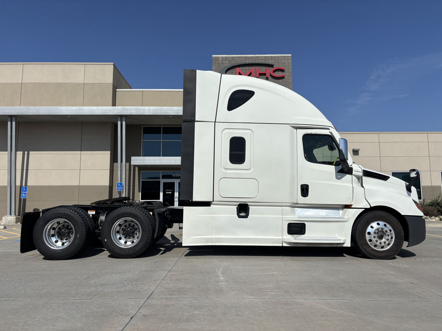 Truck side view of a white Freightliner PT126064ST conventional sleeper tractor in a parking lot with a building in the background.