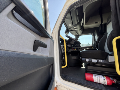 Cab interior of a Freightliner truck with open door, driver's seat visible, steering wheel, dashboard, and a red fire extinguisher on the floor.