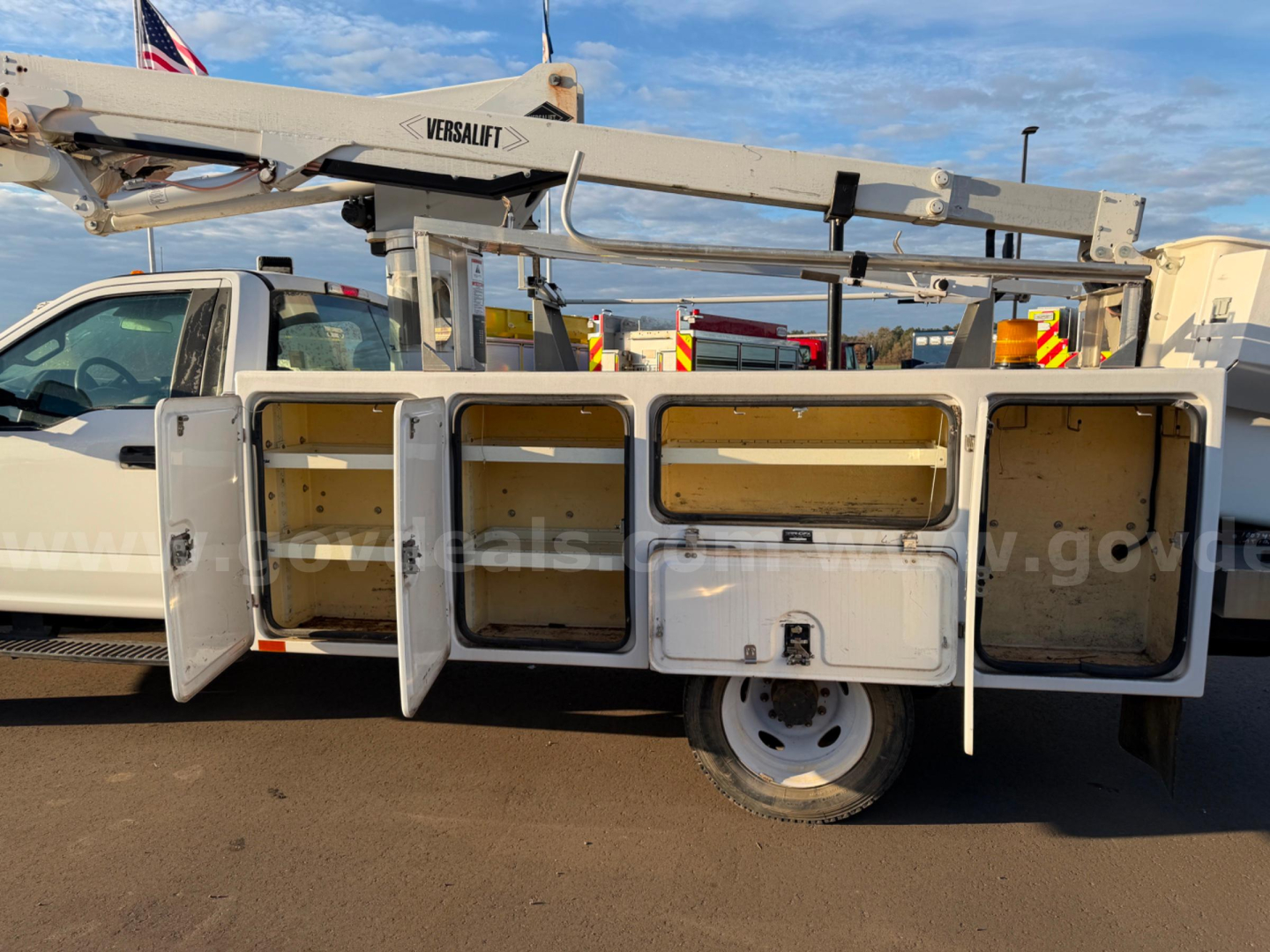 Truck bed utility compartments with open doors on a bucket truck body, Versalift lift arm overhead, white cab, desert ground.