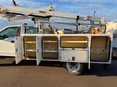 Truck bed utility compartments with open doors on a bucket truck body, Versalift lift arm overhead, white cab, desert ground.