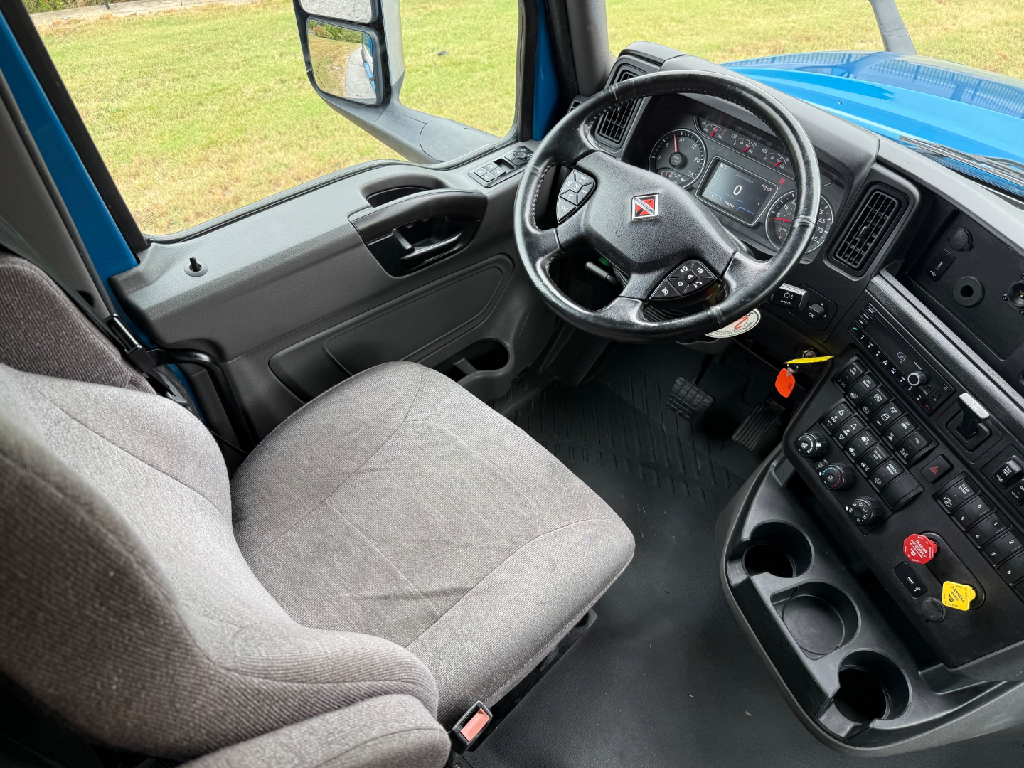 Truck cab interior showing steering wheel with International logo, instrument cluster, dashboard controls, and grey driver seat. Center console with multiple switches and cup holders.
