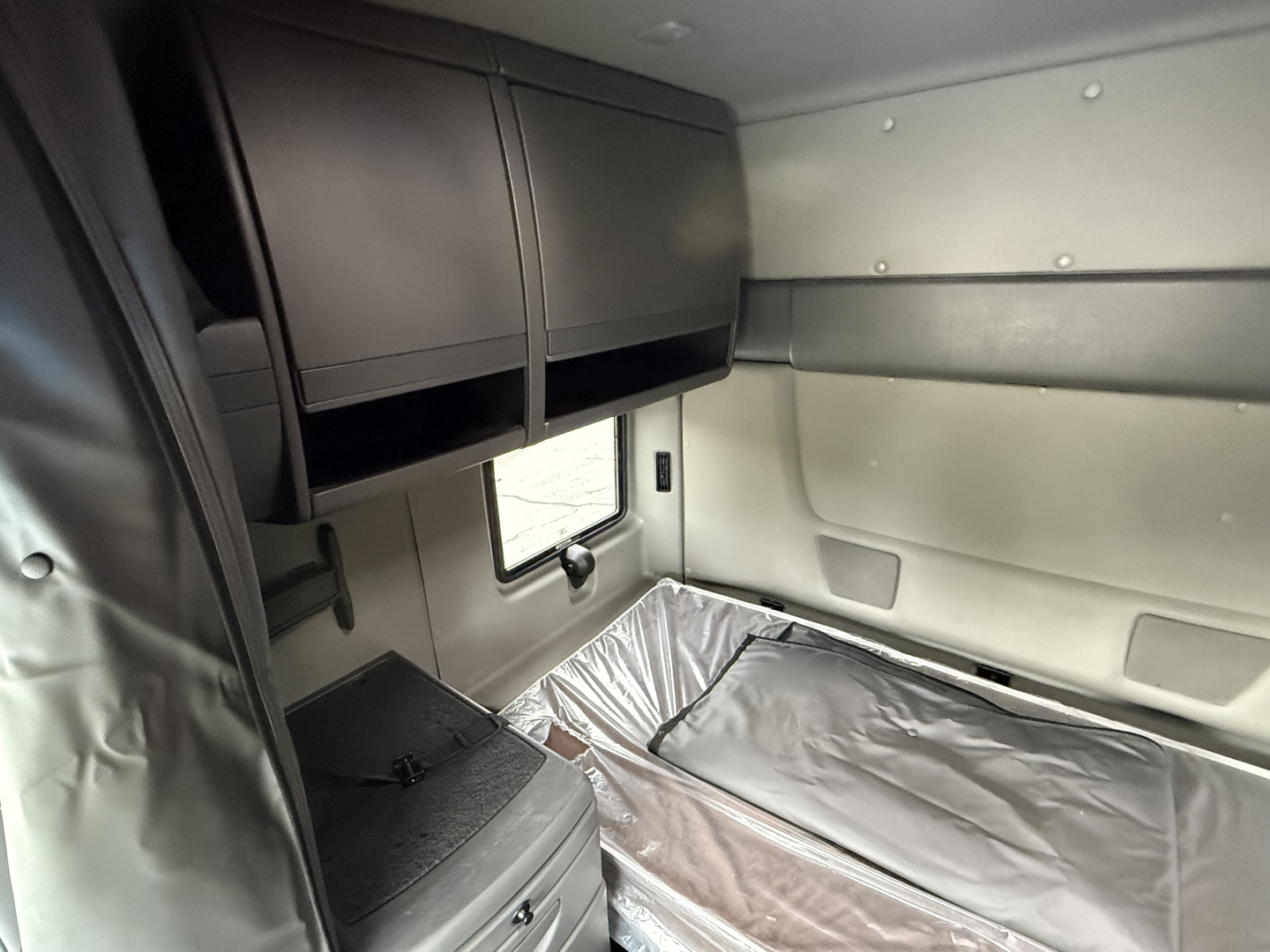 Interior sleeper cab of a 2024 International LT625 truck showing overhead storage cabinets, window, and sleeping berth with protective cover.