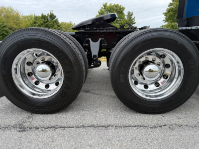 Dual rear wheels of a tractor truck with chrome hubcaps and black tires, trailer chassis and fifth wheel visible in background.