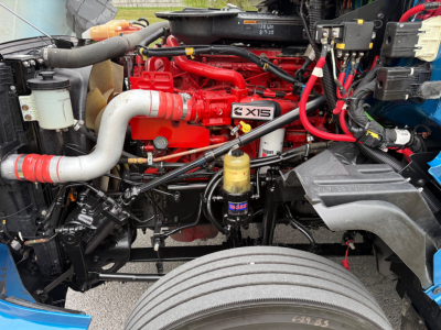 Truck engine bay with red engine block and CX15 badge, various hoses, filters, wiring harnesses, and a large front tire visible.