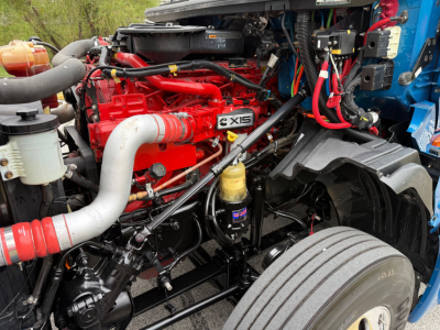 Truck engine bay with red Cummins X15 engine and surrounding components in blue-wheeled chassis, showing hoses, wiring, and air intake.
