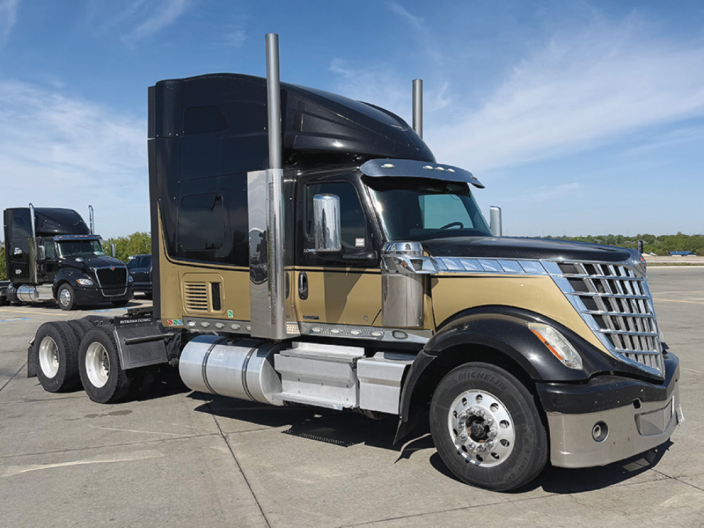 Truck side view showing 2022 International LoneStar conventional sleeper with gold/brown paint, dual exhaust stacks, chrome bumper and wheels.