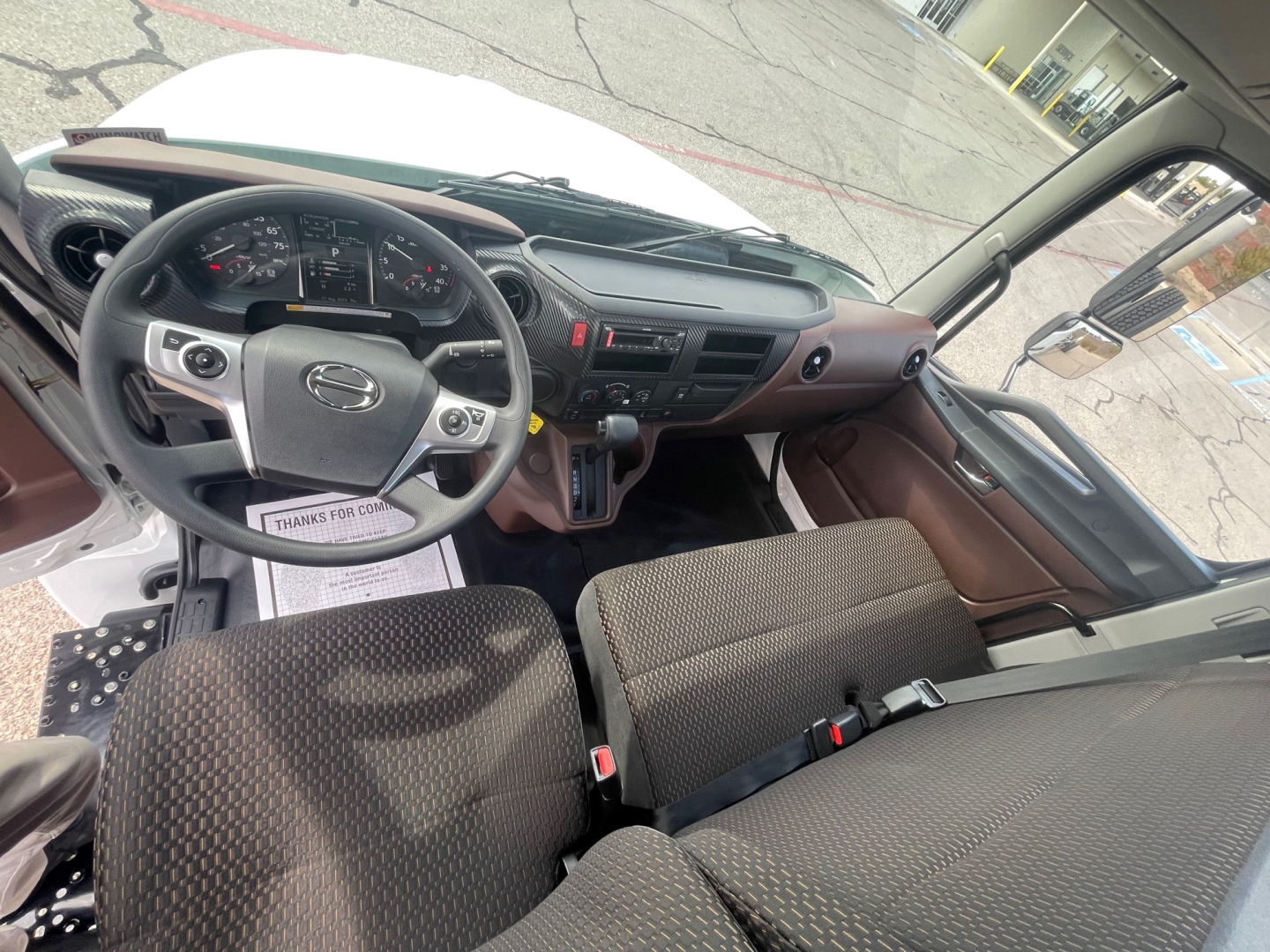 Cab interior of 2025 Hino L6 showing steering wheel with controls, dashboard, and front bench seat; driver-side door with window controls visible.