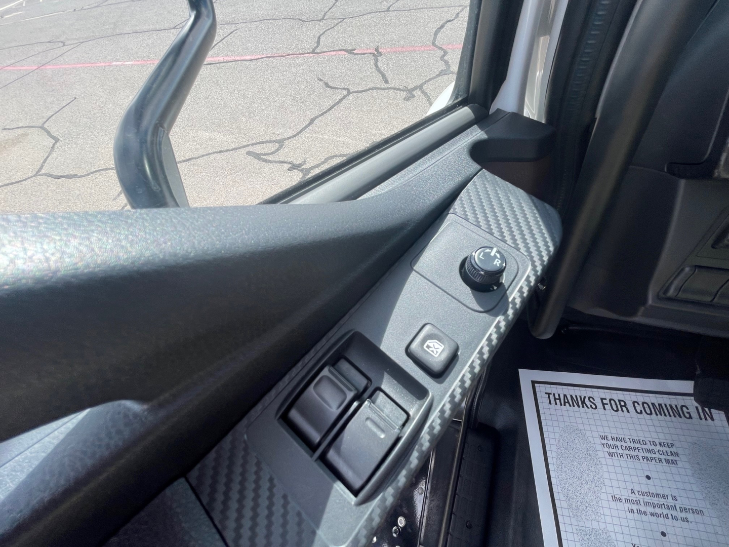 Cab interior view of a 2025 Hino L6 truck showing door panel with mirror switch, window controls, and carbon-fiber style trim.