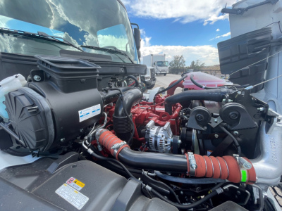 Truck engine bay of a 2025 Hino L6; visible components include radiator hoses, turbo intake, alternator, belts, coolant reservoir, and various hoses and wiring in an open engine compartment.