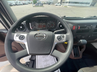 Vehicle interior steering wheel and dashboard of a 2025 Hino L6. Steering wheel with Hino logo, instrument cluster, center console, and gear shifter visible.