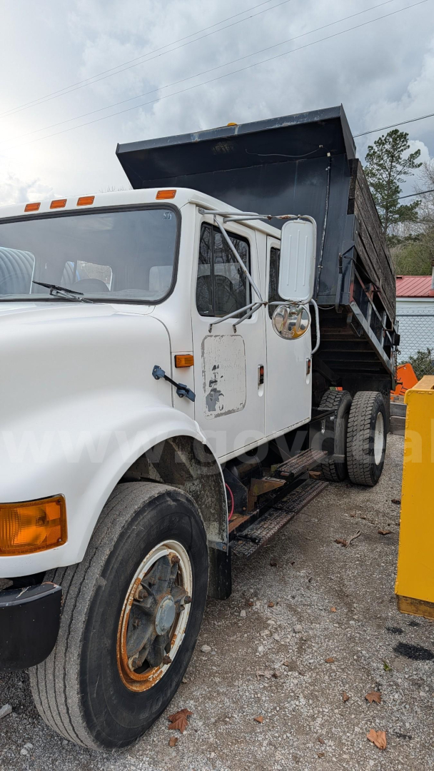 White 1990 International 4700 dump truck side view showing dump bed raised and front wheel, cab with orange turn signals and side mirror.