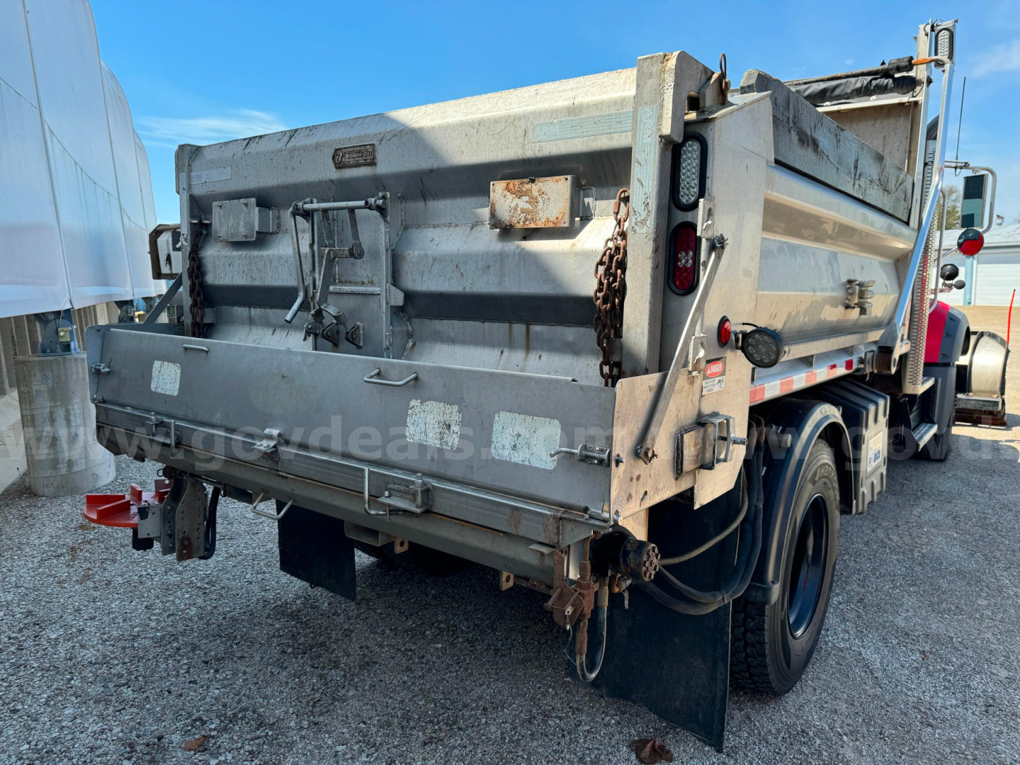 Rear/side view of a 2019 Mack Granite dump truck with plow equipment, metal tailgate, chains, and reflective safety lights. Side and rear view show dual rear wheels and mud flaps.