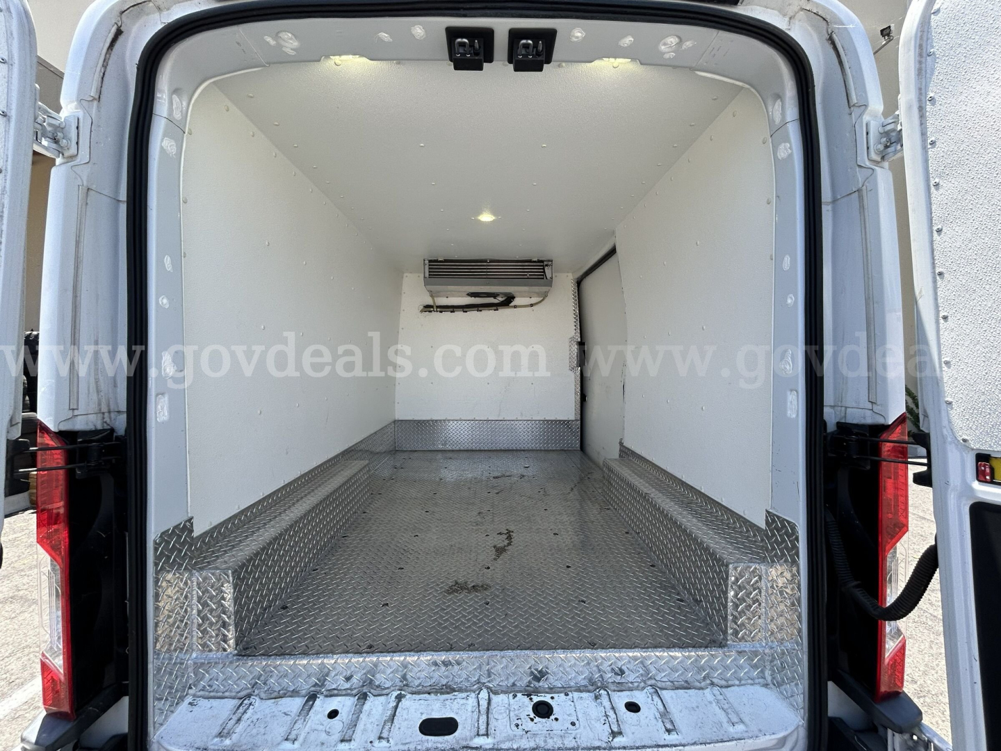 Ford Transit refrigerated cargo van with open rear doors showing empty cargo area, white insulated walls, metal diamond plate floor and ceiling-mounted evaporator unit.