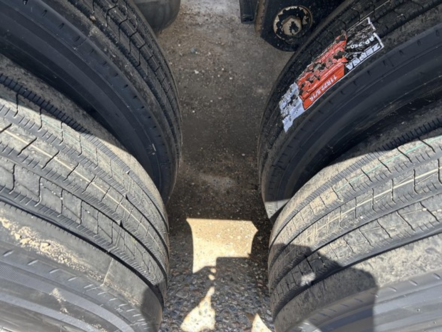 Twin rear axles of a dump trailer with tires and a small red/white sticker on one tire; visible dirt and gravel on the ground.