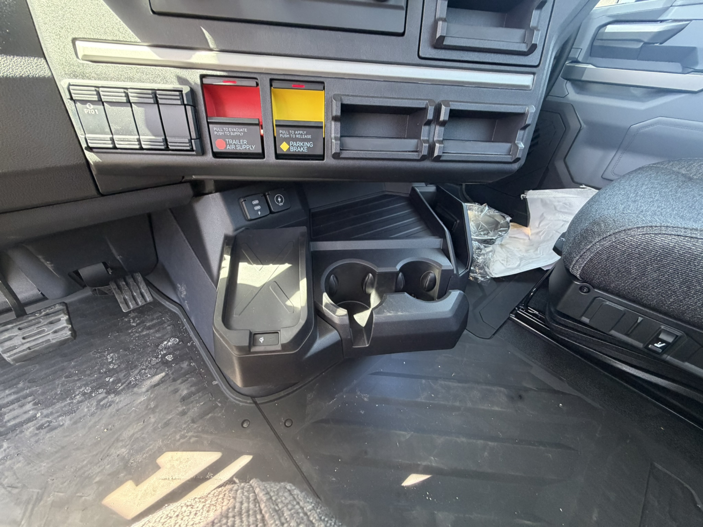 Interior cab area showing dashboard switches and cup holders in a Mack AN64T truck cab, with pedals and floor mat visible.
