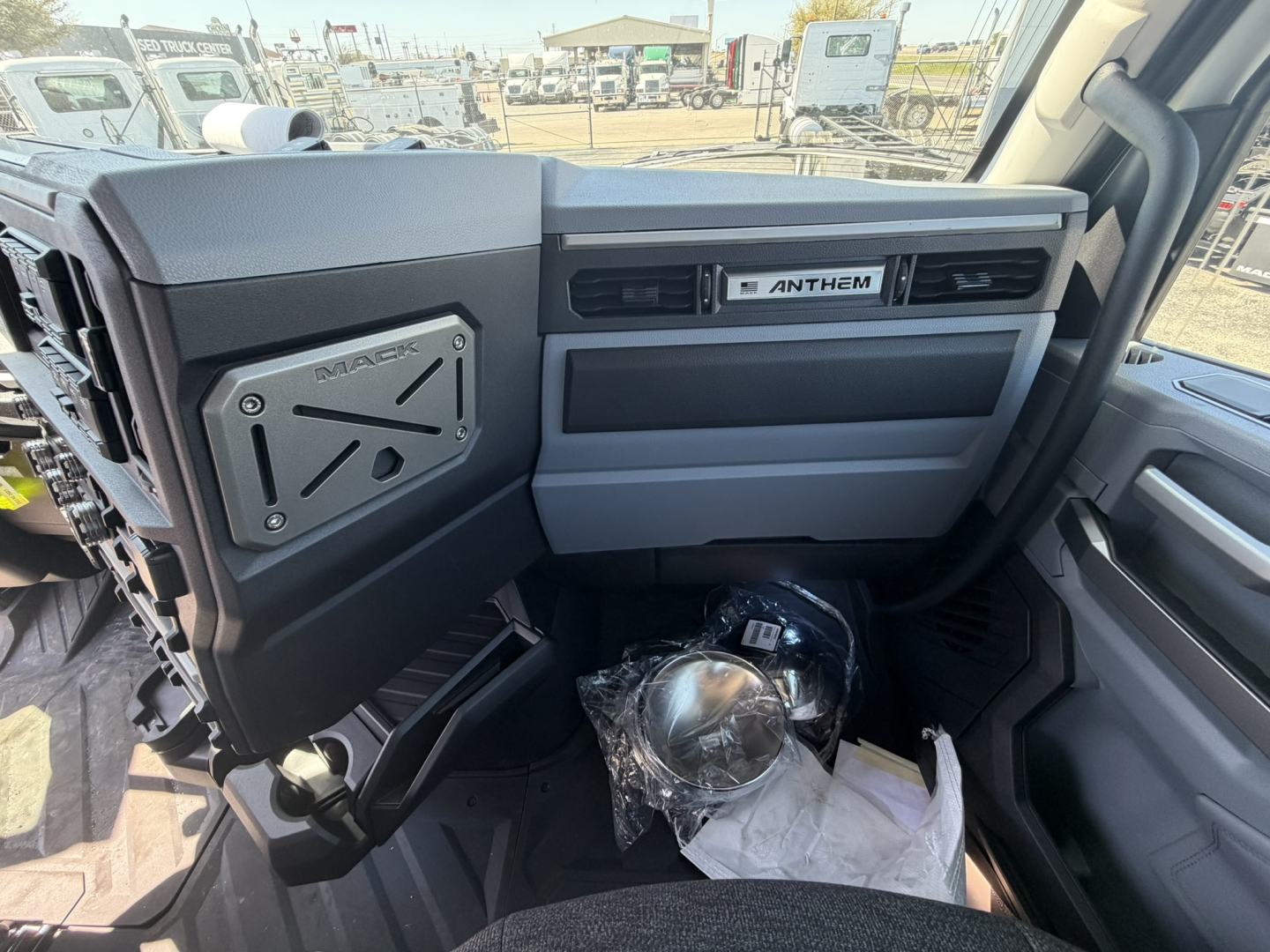 Truck cockpit interior showing Mack ANTHEM badge on dashboard, glove compartment, and steering area; view from passenger side with plastic-wrapped item on floor.