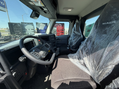 Front cab interior of a 2027 Mack AN64T daycab showing steering wheel, driver's seat wrapped in plastic, dashboard, and passenger seat.