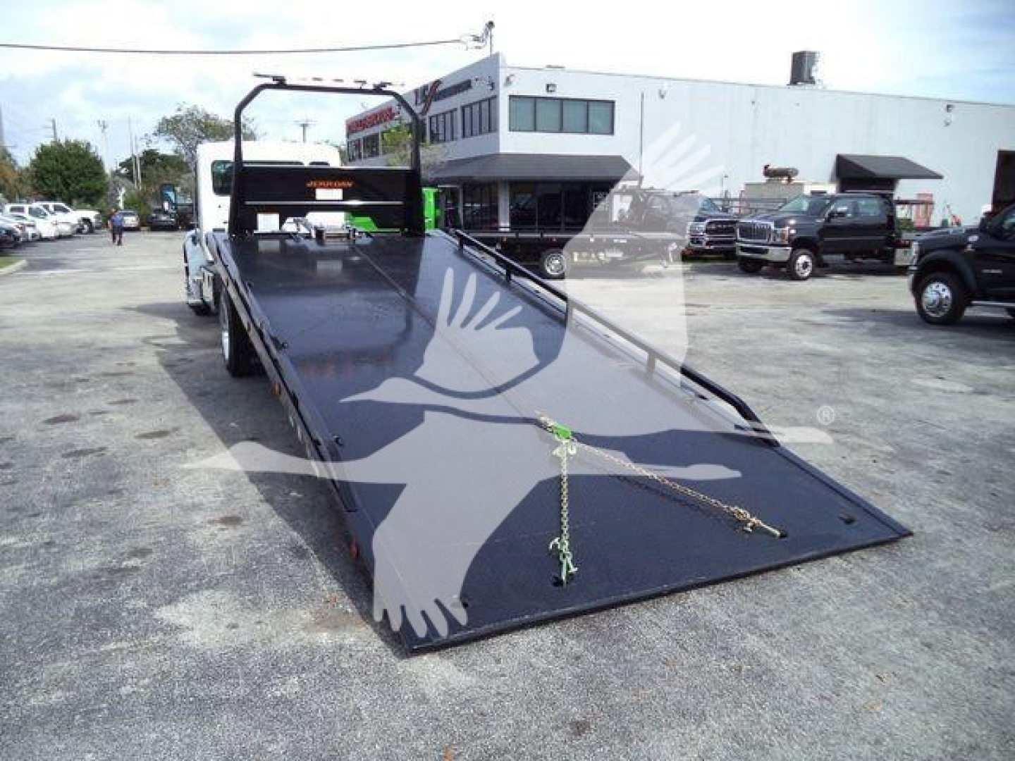 Kenworth T280 rollback truck flatbed with ramp in lowered position, parked lot with building in background. Visible chains and hooks on deck.